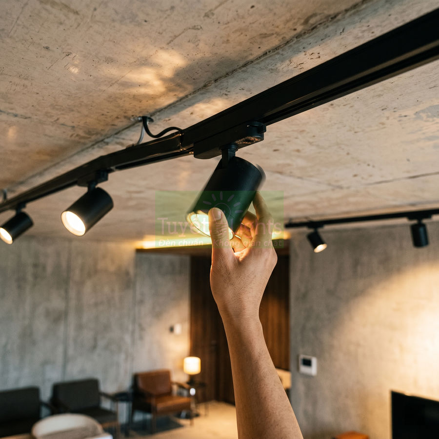 Đèn rọi ray trần nhà màu đen trong phòng khách hiện đại, tay người đang điều chỉnh hướng chiếu sáng