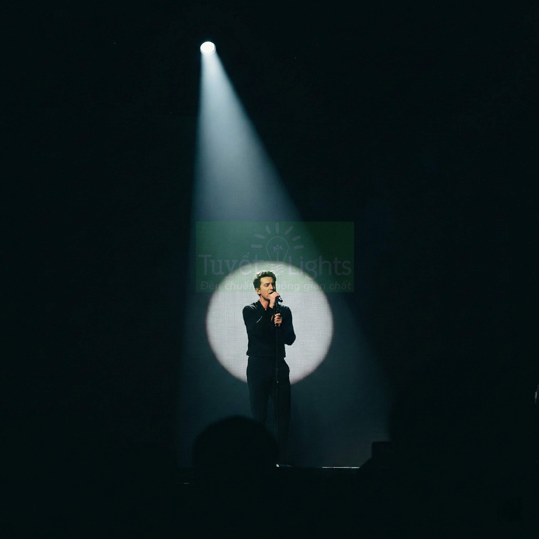 Ca sĩ đứng trên sân khấu dưới ánh đèn sân khấu tập trung, hát vào micro trên chân đế